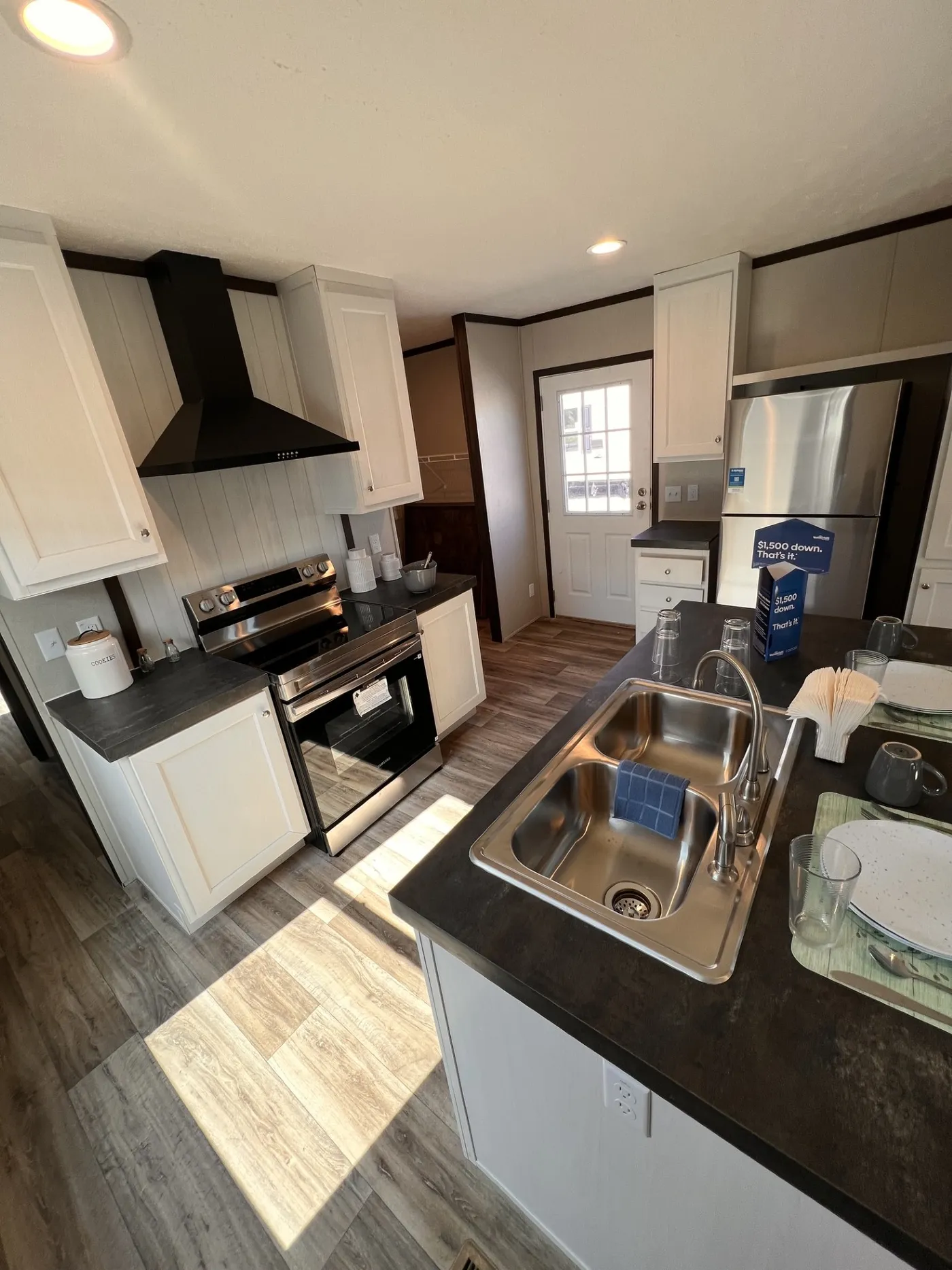 Bright modern kitchen featuring white cabinetry, dark countertops, stainless-steel appliances, and a spacious center island with a double sink. Warm wood-looking flooring and the contrasting black range hood give it a clean, stylish finish.